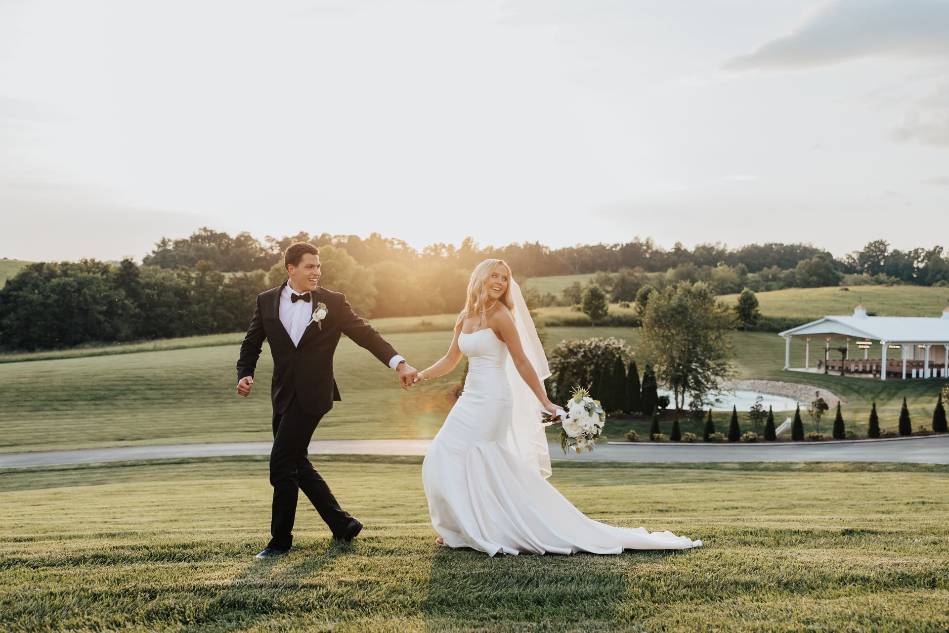 A couple in formal attire walking hand in hand across a sunlit field at golden hour